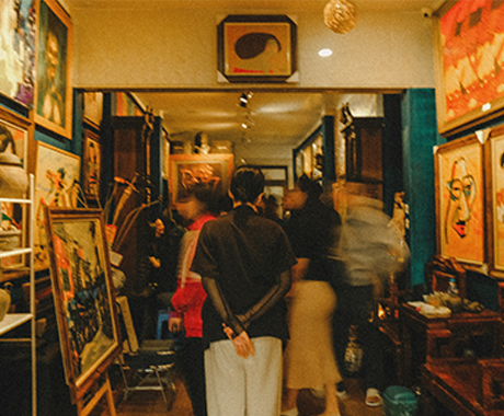 A crowded art gallery room with vibrant paintings on the walls. People are gathered, blurred in motion, creating a lively and engaging atmosphere.