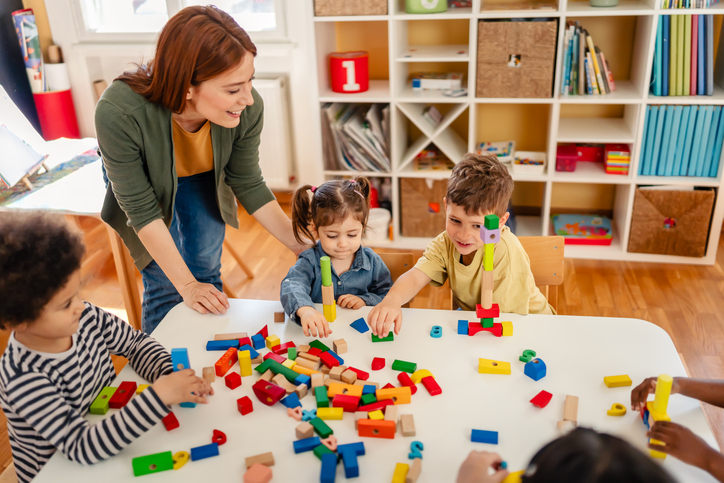 A teacher and five children play with colorful blocks around a white table in a classroom. Shelves and educational materials fill the background, creating a joyful and engaging atmosphere.