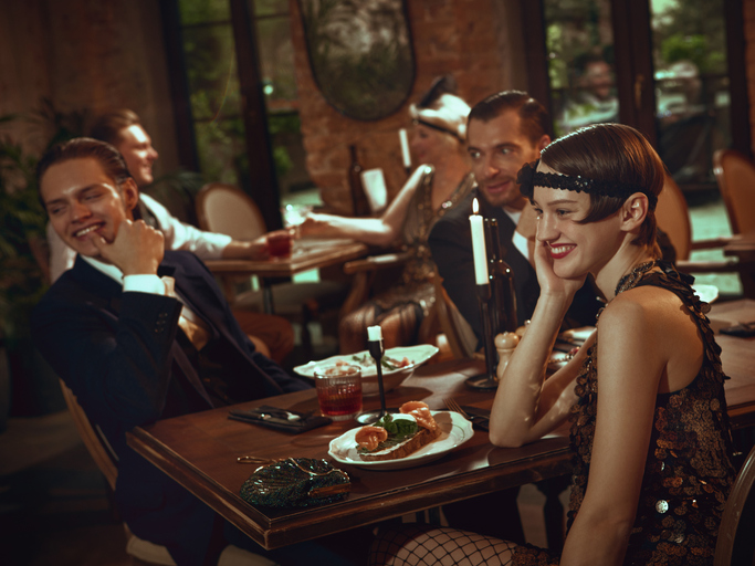 A lively 1920s-themed party scene with elegantly dressed guests laughing around a dimly lit table set with candles, cocktails, and appetizers.
