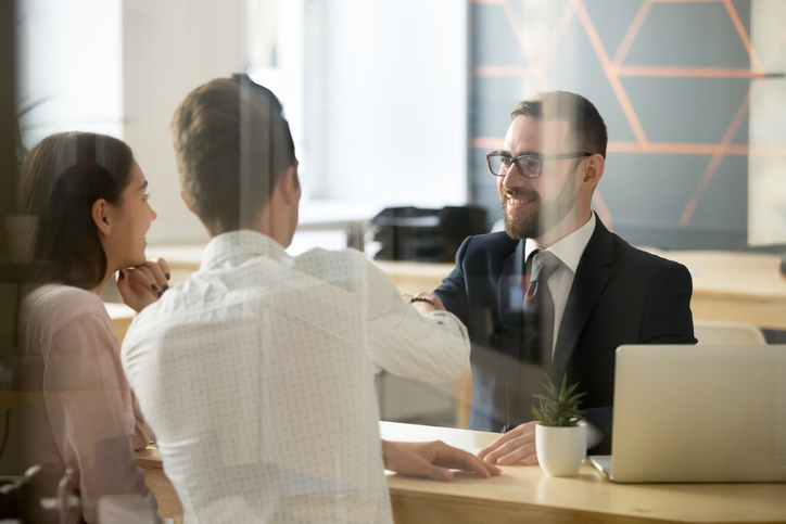 A business professional in a suit smiles confidently while shaking hands with a client couple across a desk, conveying trust and agreement in a modern office.