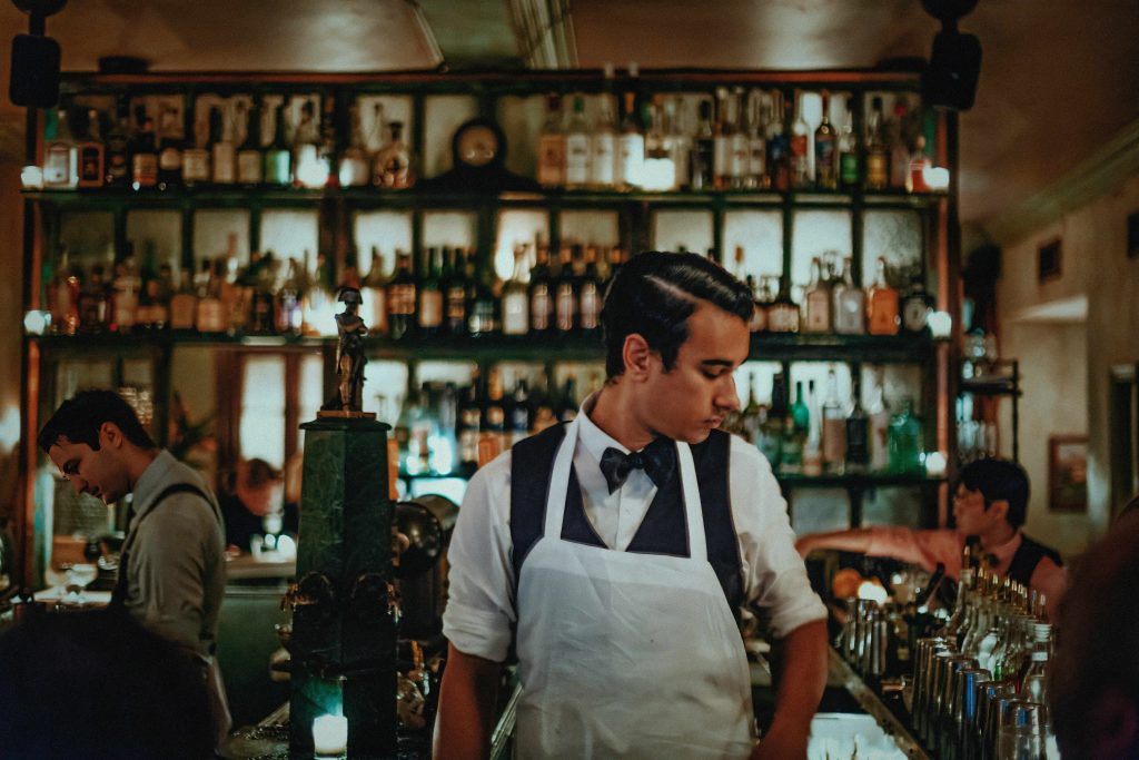 Bartender in a bow tie and apron mixes drinks at a dimly lit, vintage-style bar, with shelves full of bottles in the background, creating a nostalgic ambiance.