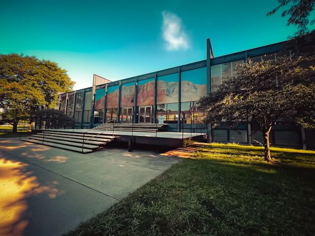 A modern glass building with large windows reflecting the sky and trees, surrounded by greenery. The scene is tranquil, bathed in warm sunlight.