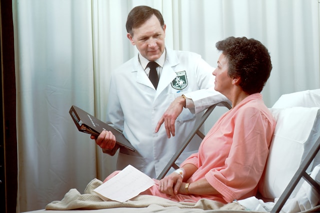 A doctor in a white coat discusses with a patient in a hospital bed, holding a clipboard. The scene conveys a caring and professional tone.